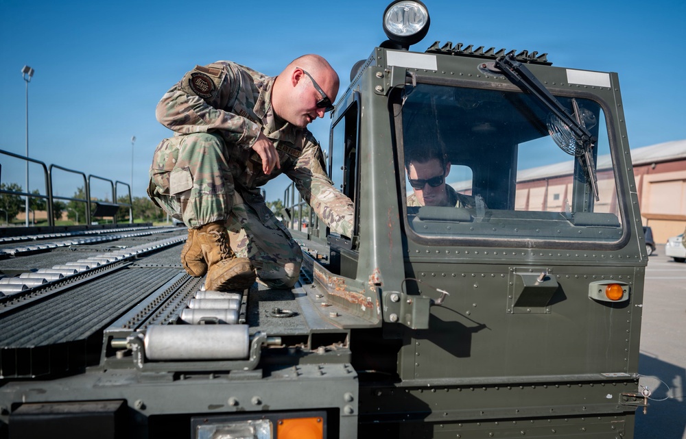 Two military personnel in OCP (Operational Camouflage Pattern) uniforms are working with a military ground support vehicle, likely a cargo loader or conveyor equipment used on flight lines. One service member is crouching on top of the roller conveyor bed inspecting or operating a component, while a second is seated inside the enclosed cab of the olive-drab vehicle. The setting appears to be a military airfield or base ramp area.