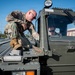 Two military personnel in OCP (Operational Camouflage Pattern) uniforms are working with a military ground support vehicle, likely a cargo loader or conveyor equipment used on flight lines. One service member is crouching on top of the roller conveyor bed inspecting or operating a component, while a second is seated inside the enclosed cab of the olive-drab vehicle. The setting appears to be a military airfield or base ramp area.