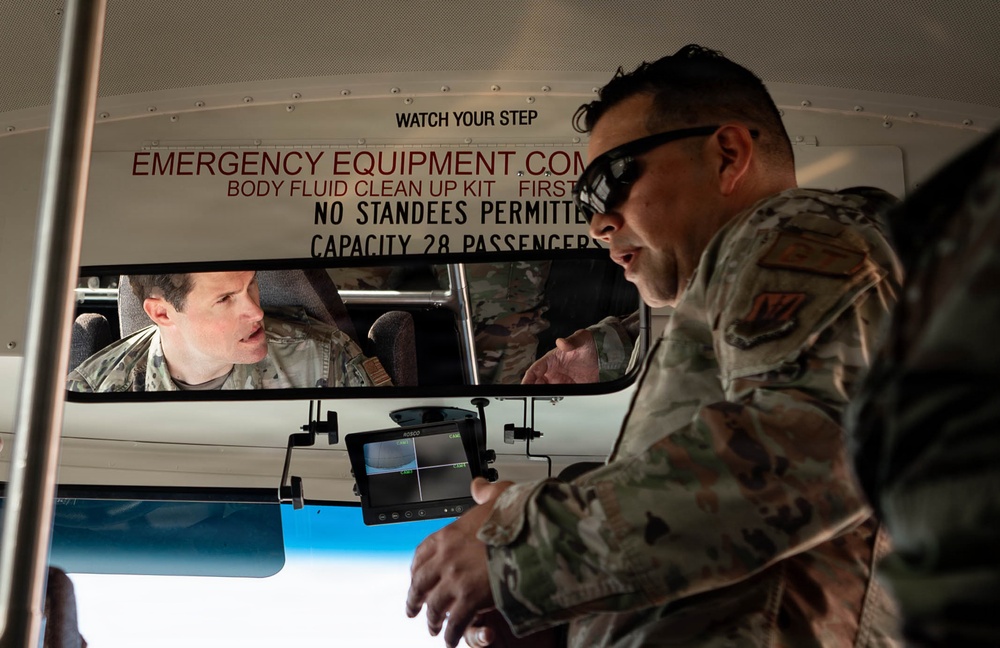 Two military personnel in camouflage uniforms are interacting inside or near a bus. One individual is seated in the driver's area while another, wearing sunglasses, stands at the front of the bus examining a small monitor device. The scene appears to involve an inspection or briefing related to bus safety or surveillance equipment.