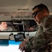Two military personnel in camouflage uniforms are interacting inside or near a bus. One individual is seated in the driver's area while another, wearing sunglasses, stands at the front of the bus examining a small monitor device. The scene appears to involve an inspection or briefing related to bus safety or surveillance equipment.