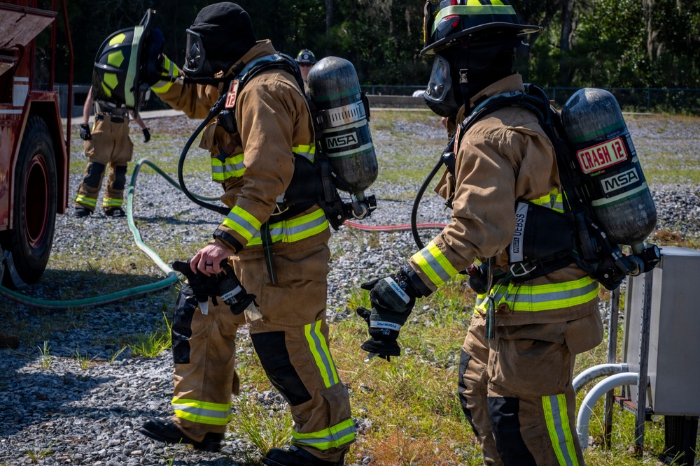 Three firefighters in full turnout gear are visible at what appears to be a training exercise or active incident scene on a gravel surface. They are equipped with MSA self-contained breathing apparatus (SCBA) and are holding breathing masks, suggesting they are preparing to enter or have just exited a hazardous environment. A fire truck is partially visible on the left, and fire hoses are laid out on the ground.