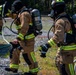 Three firefighters in full turnout gear are visible at what appears to be a training exercise or active incident scene on a gravel surface. They are equipped with MSA self-contained breathing apparatus (SCBA) and are holding breathing masks, suggesting they are preparing to enter or have just exited a hazardous environment. A fire truck is partially visible on the left, and fire hoses are laid out on the ground.