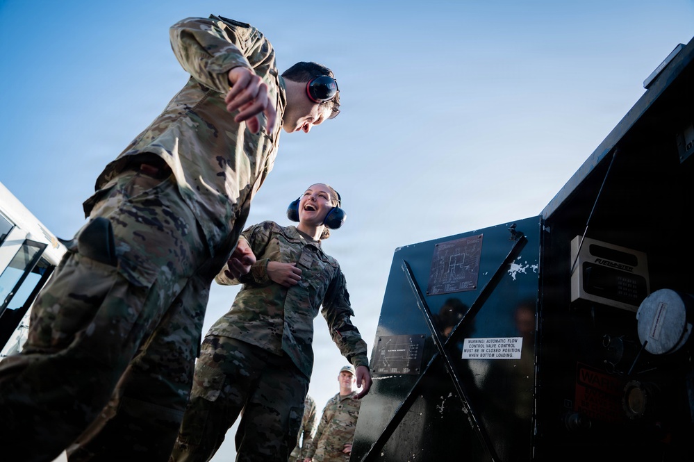 Two military personnel in OCP (Operational Camouflage Pattern) uniforms are laughing and interacting near what appears to be a fuel truck or ground support equipment on a flight line. Both individuals are wearing hearing protection (ear muffs), suggesting proximity to loud aircraft or machinery. A third service member is visible in the background wearing a cap.