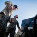 Two military personnel in OCP (Operational Camouflage Pattern) uniforms are laughing and interacting near what appears to be a fuel truck or ground support equipment on a flight line. Both individuals are wearing hearing protection (ear muffs), suggesting proximity to loud aircraft or machinery. A third service member is visible in the background wearing a cap.