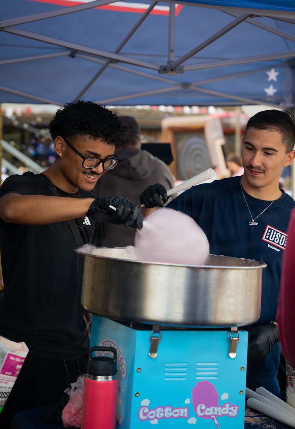 Two young men are operating a cotton candy machine at what appears to be a USO volunteer event. The person in the foreground, wearing black gloves and glasses, is actively spinning cotton candy on a cone while smiling. They are working under a blue canopy tent with stars and stripes decorations, suggesting a patriotic or military-affiliated event.