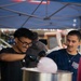 Two young men are operating a cotton candy machine at what appears to be a USO volunteer event. The person in the foreground, wearing black gloves and glasses, is actively spinning cotton candy on a cone while smiling. They are working under a blue canopy tent with stars and stripes decorations, suggesting a patriotic or military-affiliated event.