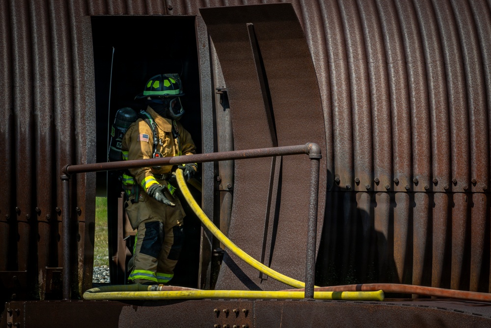 A firefighter in full personal protective equipment is positioned in the doorway of a corrugated metal structure, likely a training prop or decommissioned rail car. The firefighter is carrying a yellow fire hose and wearing full turnout gear including SCBA (self-contained breathing apparatus). This appears to be a firefighter training exercise based on the controlled setting and equipment.
