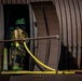 A firefighter in full personal protective equipment is positioned in the doorway of a corrugated metal structure, likely a training prop or decommissioned rail car. The firefighter is carrying a yellow fire hose and wearing full turnout gear including SCBA (self-contained breathing apparatus). This appears to be a firefighter training exercise based on the controlled setting and equipment.
