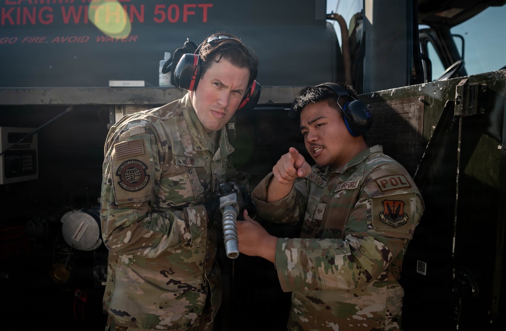 Two U.S. Air Force personnel in OCP (Operational Camouflage Pattern) uniforms are engaged in what appears to be a training or operational briefing near a fuel truck. The airman on the right, wearing a 'POL' (Petroleum, Oils, and Lubricants) patch and blue ear protection, is pointing and explaining something while holding a fuel nozzle/coupling. Both individuals are wearing hearing protection, suggesting a noisy flight line environment.