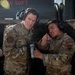 Two U.S. Air Force personnel in OCP (Operational Camouflage Pattern) uniforms are engaged in what appears to be a training or operational briefing near a fuel truck. The airman on the right, wearing a 'POL' (Petroleum, Oils, and Lubricants) patch and blue ear protection, is pointing and explaining something while holding a fuel nozzle/coupling. Both individuals are wearing hearing protection, suggesting a noisy flight line environment.