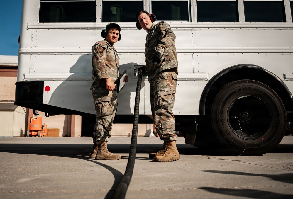 Two military personnel in OCP (Operational Camouflage Pattern) uniforms are standing near a white bus on what appears to be a military installation or airfield. Both individuals are wearing hearing protection (ear muffs) and tan combat boots, suggesting proximity to loud equipment or aircraft. One person is holding a large black hose or cable while appearing to communicate with the other.