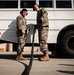 Two military personnel in OCP (Operational Camouflage Pattern) uniforms are standing near a white bus on what appears to be a military installation or airfield. Both individuals are wearing hearing protection (ear muffs) and tan combat boots, suggesting proximity to loud equipment or aircraft. One person is holding a large black hose or cable while appearing to communicate with the other.