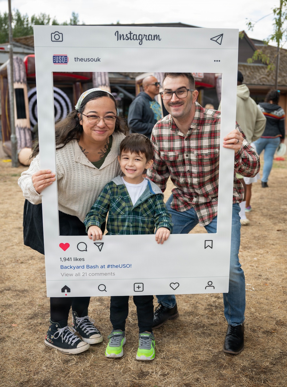 A family of three — two adults and a young child — poses for a photo at an outdoor event using a large prop Instagram frame. The frame displays the USO UK account (@theusouk) with the caption 'Backyard Bash at #theUSO!' and shows 1,941 likes. The setting appears to be a casual outdoor gathering or festival with other attendees visible in the background.