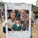 A family of three — two adults and a young child — poses for a photo at an outdoor event using a large prop Instagram frame. The frame displays the USO UK account (@theusouk) with the caption 'Backyard Bash at #theUSO!' and shows 1,941 likes. The setting appears to be a casual outdoor gathering or festival with other attendees visible in the background.
