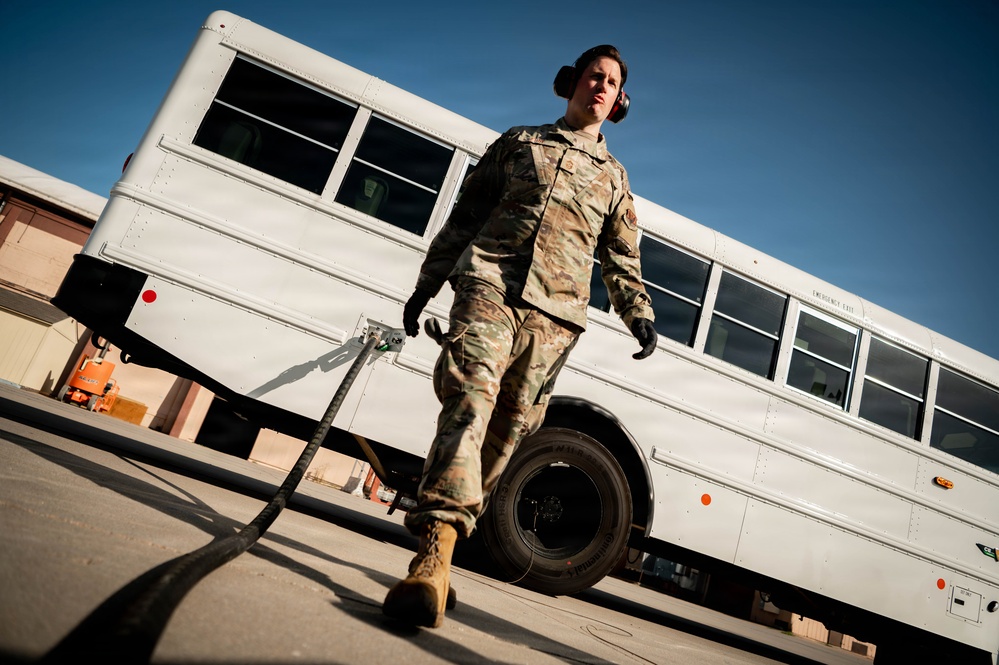 A military service member in OCP (Operational Camouflage Pattern) uniform is walking near a large white bus on what appears to be a military installation or airfield. The individual is wearing hearing protection and black gloves while dragging or carrying a cable or hose. The low-angle photograph creates a dramatic perspective emphasizing the subject against a clear blue sky.