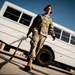 A military service member in OCP (Operational Camouflage Pattern) uniform is walking near a large white bus on what appears to be a military installation or airfield. The individual is wearing hearing protection and black gloves while dragging or carrying a cable or hose. The low-angle photograph creates a dramatic perspective emphasizing the subject against a clear blue sky.
