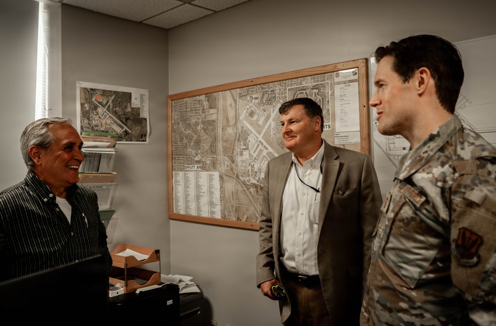 Three men are engaged in conversation inside what appears to be a military office or administrative workspace. One man is seated at a desk, one is standing in civilian business attire, and the third is wearing a military camouflage uniform. A map labeled 'Offutt AFB' is prominently displayed on the wall behind them.
