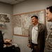 Three men are engaged in conversation inside what appears to be a military office or administrative workspace. One man is seated at a desk, one is standing in civilian business attire, and the third is wearing a military camouflage uniform. A map labeled 'Offutt AFB' is prominently displayed on the wall behind them.