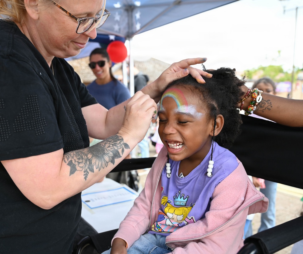 A face painter at an outdoor community event is applying a rainbow design to a young girl's forehead. The child is smiling broadly and appears delighted by the experience. The setting appears to be a festive outdoor fair or community gathering with a tent canopy visible in the background.