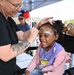 A face painter at an outdoor community event is applying a rainbow design to a young girl's forehead. The child is smiling broadly and appears delighted by the experience. The setting appears to be a festive outdoor fair or community gathering with a tent canopy visible in the background.