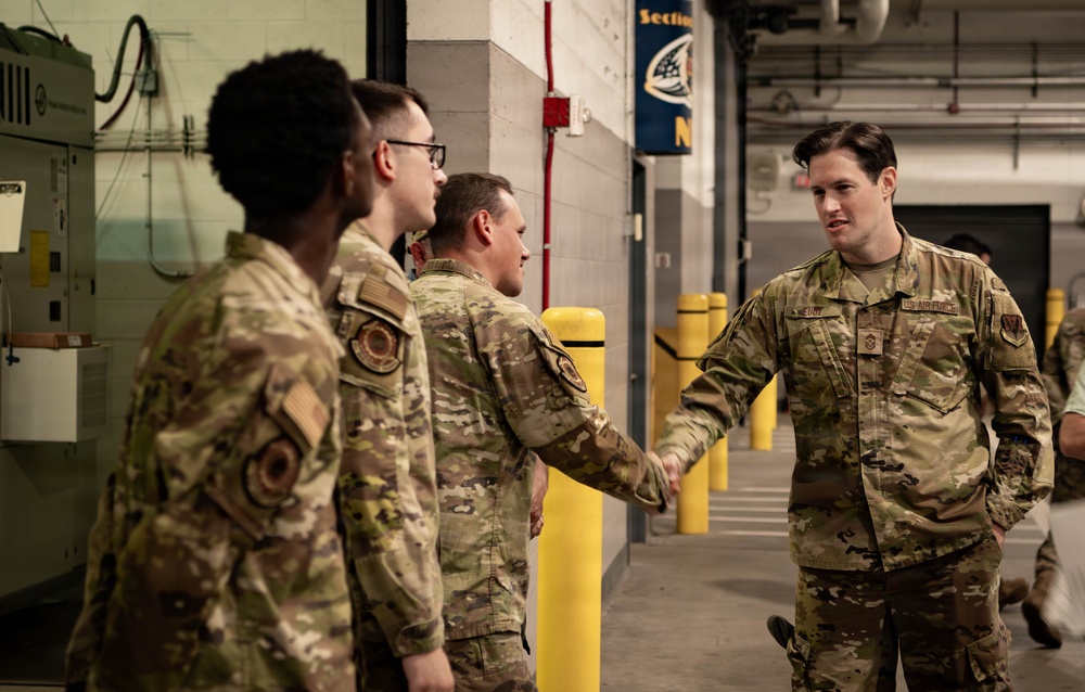 A group of U.S. Air Force personnel in OCP (Operational Camouflage Pattern) uniforms are gathered in what appears to be an indoor facility, possibly a hangar or arena corridor. One airman on the right is shaking hands with another, suggesting a greeting or recognition event. The setting includes industrial infrastructure and what appears to be a sports or event venue banner in the background.