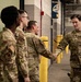 A group of U.S. Air Force personnel in OCP (Operational Camouflage Pattern) uniforms are gathered in what appears to be an indoor facility, possibly a hangar or arena corridor. One airman on the right is shaking hands with another, suggesting a greeting or recognition event. The setting includes industrial infrastructure and what appears to be a sports or event venue banner in the background.