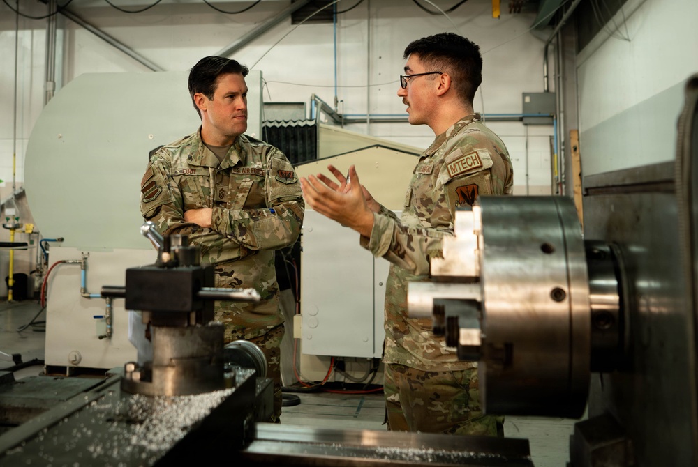 Two U.S. Air Force personnel in OCP (Operational Camouflage Pattern) uniforms are engaged in a discussion inside what appears to be a machine shop or maintenance facility. The individual on the right, whose name tape reads 'MTECH', is holding a document or folder and appears to be briefing or explaining something to the senior airman on the left. Industrial machining equipment, including a lathe, is visible in the foreground.