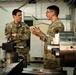 Two U.S. Air Force personnel in OCP (Operational Camouflage Pattern) uniforms are engaged in a discussion inside what appears to be a machine shop or maintenance facility. The individual on the right, whose name tape reads 'MTECH', is holding a document or folder and appears to be briefing or explaining something to the senior airman on the left. Industrial machining equipment, including a lathe, is visible in the foreground.