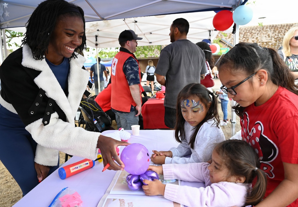 A community outdoor event featuring a balloon art activity station where a smiling adult woman is presenting a purple balloon octopus sculpture to three young girls. One child has decorative face paint with a butterfly/fantasy design. In the background, volunteers in American Red Cross vests are visible at adjacent tables.