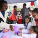 A community outdoor event featuring a balloon art activity station where a smiling adult woman is presenting a purple balloon octopus sculpture to three young girls. One child has decorative face paint with a butterfly/fantasy design. In the background, volunteers in American Red Cross vests are visible at adjacent tables.