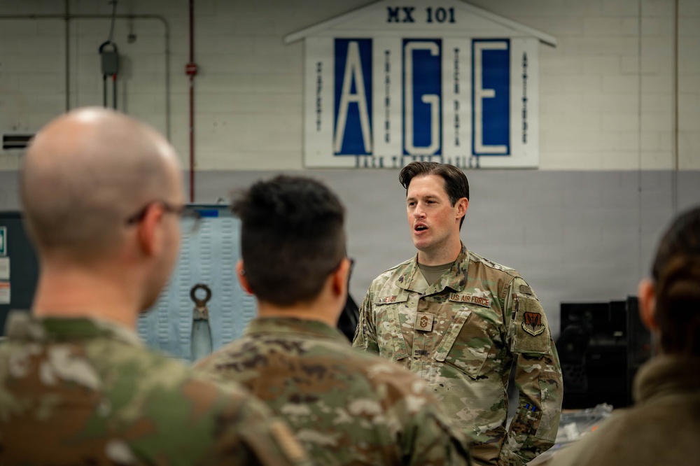 A US Air Force personnel member in OCP (Operational Camouflage Pattern) uniform is addressing a group of fellow airmen inside a maintenance facility. The speaker appears to be a senior non-commissioned officer conducting a briefing or training session. The setting is identified as an AGE (Aerospace Ground Equipment) shop, designated MX 101.