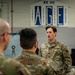 A US Air Force personnel member in OCP (Operational Camouflage Pattern) uniform is addressing a group of fellow airmen inside a maintenance facility. The speaker appears to be a senior non-commissioned officer conducting a briefing or training session. The setting is identified as an AGE (Aerospace Ground Equipment) shop, designated MX 101.