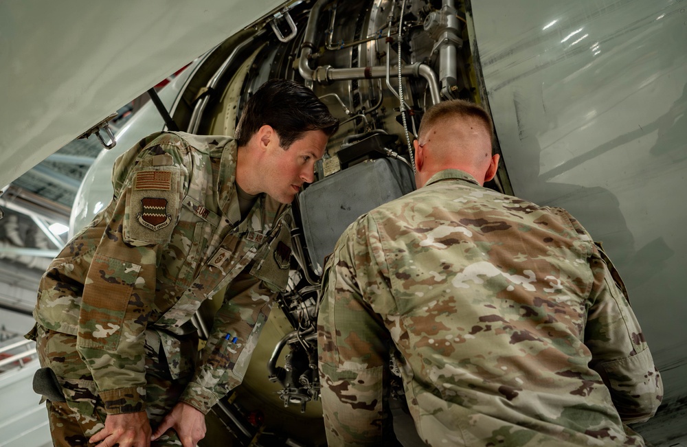 Two U.S. Air Force personnel in OCP (Operational Camouflage Pattern) uniforms are inspecting or performing maintenance on an aircraft component, likely inside a hangar. The open access panel reveals internal mechanical components including tubing, hydraulic lines, and structural elements. The interaction suggests a supervisory or instructional exchange between the two individuals.
