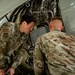 Two U.S. Air Force personnel in OCP (Operational Camouflage Pattern) uniforms are inspecting or performing maintenance on an aircraft component, likely inside a hangar. The open access panel reveals internal mechanical components including tubing, hydraulic lines, and structural elements. The interaction suggests a supervisory or instructional exchange between the two individuals.
