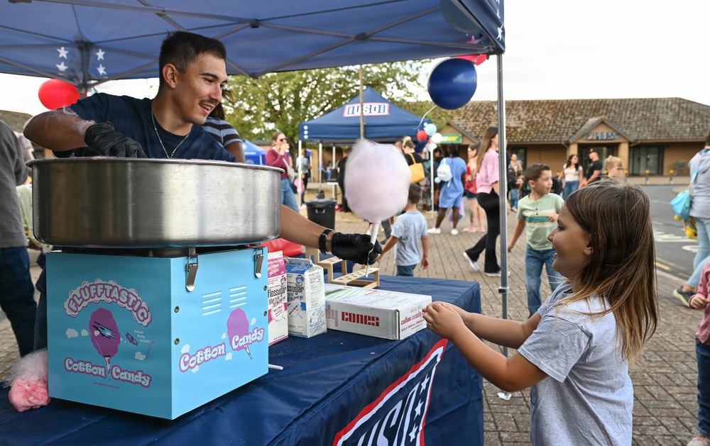 A young man operates a 'Flufftastic Cotton Candy' booth at an outdoor fair or community event, handing a pink cotton candy to a young girl. The scene takes place under a blue canopy tent decorated with stars, surrounded by a crowd of people in the background. The atmosphere is festive and family-friendly, with red, white, and blue balloons visible overhead.