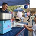 A young man operates a 'Flufftastic Cotton Candy' booth at an outdoor fair or community event, handing a pink cotton candy to a young girl. The scene takes place under a blue canopy tent decorated with stars, surrounded by a crowd of people in the background. The atmosphere is festive and family-friendly, with red, white, and blue balloons visible overhead.