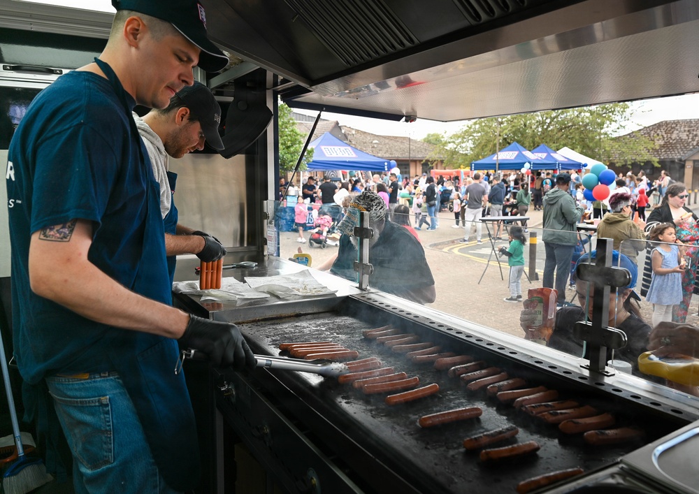 Two food truck workers wearing black gloves and baseball caps are grilling a large number of hot dogs/sausages on a flat-top griddle. The scene is set at an outdoor community event or festival, visible through the food truck's service window, with crowds of people, blue vendor tents, and balloons in the background. The workers appear to be actively managing the grill during a busy service period.