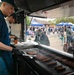 Two food truck workers wearing black gloves and baseball caps are grilling a large number of hot dogs/sausages on a flat-top griddle. The scene is set at an outdoor community event or festival, visible through the food truck's service window, with crowds of people, blue vendor tents, and balloons in the background. The workers appear to be actively managing the grill during a busy service period.