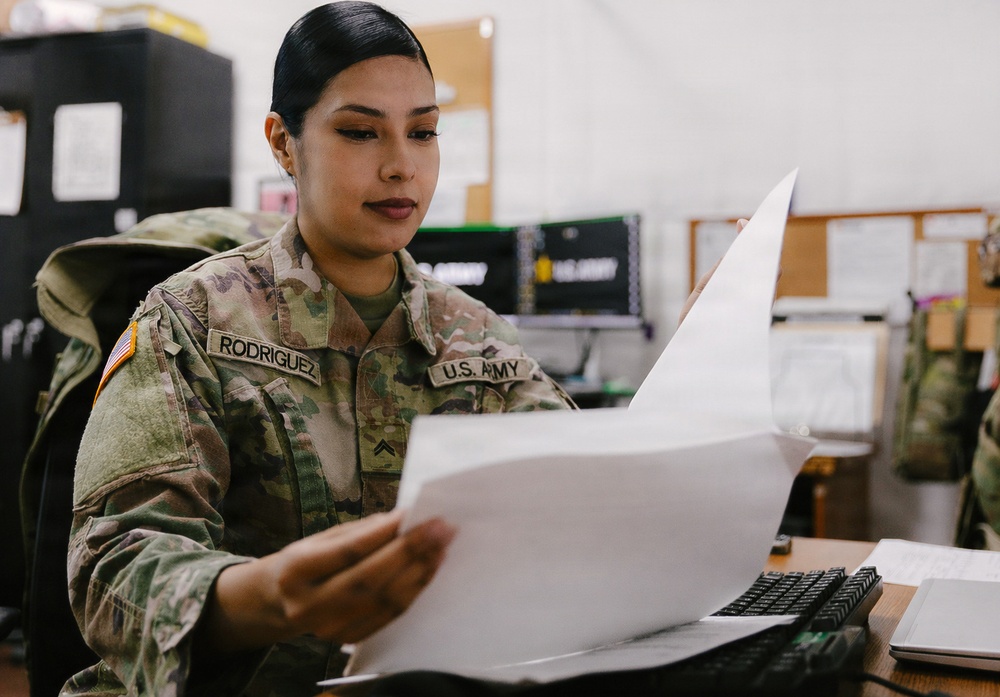 A U.S. Army soldier named Rodriguez, wearing Operational Camouflage Pattern (OCP) uniform, is seated at a desk reviewing printed documents. The setting appears to be a military administrative office environment. The soldier holds rank insignia consistent with a Private First Class or Specialist grade.