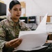 A U.S. Army soldier named Rodriguez, wearing Operational Camouflage Pattern (OCP) uniform, is seated at a desk reviewing printed documents. The setting appears to be a military administrative office environment. The soldier holds rank insignia consistent with a Private First Class or Specialist grade.