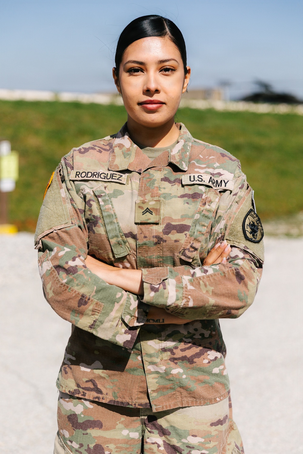 A U.S. Army soldier named Rodriguez stands at ease with arms crossed in an outdoor military setting. She is wearing the standard Army Combat Uniform (ACU) in OCP (Operational Camouflage Pattern) with visible rank insignia of Corporal. The image appears to be a formal or semi-formal portrait taken in daylight.