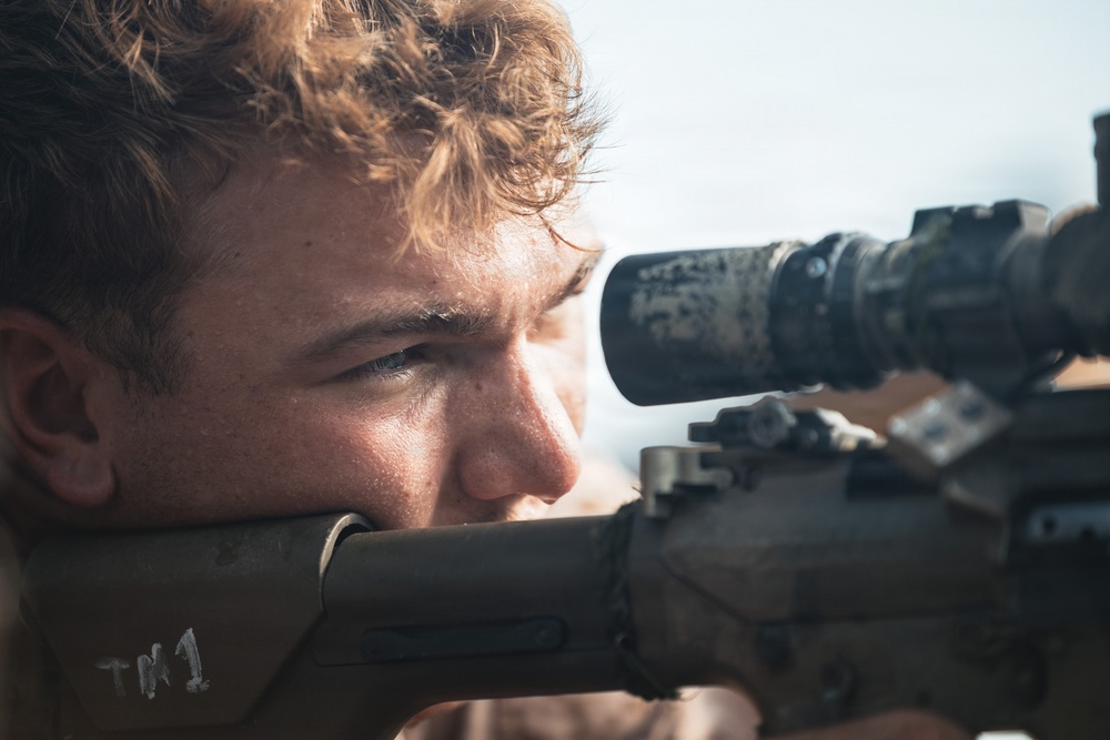A close-up photograph of a young male individual aiming through the scope of a rifle. The image is tightly framed, focusing on his face and the weapon's optic system. The subject appears focused and is in a prone or low shooting position.