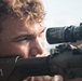 A close-up photograph of a young male individual aiming through the scope of a rifle. The image is tightly framed, focusing on his face and the weapon's optic system. The subject appears focused and is in a prone or low shooting position.