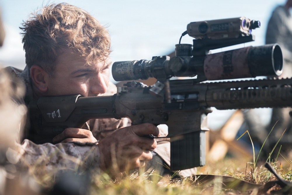A young male in camouflage military uniform is lying prone on the ground aiming a rifle equipped with multiple optical attachments including a scope and what appears to be a laser rangefinder or targeting device. The image is a close-up shot capturing the shooter's focused expression as he looks through the rifle scope. The setting appears to be an outdoor training or field environment.