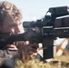 A young male in camouflage military uniform is lying prone on the ground aiming a rifle equipped with multiple optical attachments including a scope and what appears to be a laser rangefinder or targeting device. The image is a close-up shot capturing the shooter's focused expression as he looks through the rifle scope. The setting appears to be an outdoor training or field environment.
