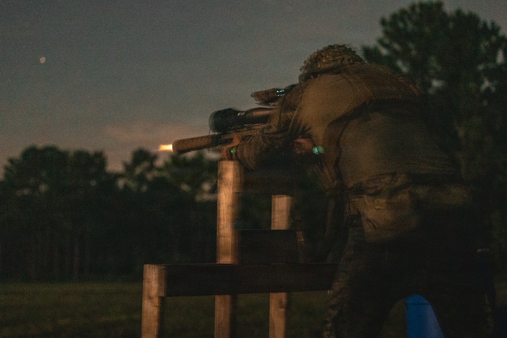 A person in military/tactical gear is firing a rifle with a suppressor from a wooden shooting bench or barricade during dusk or nighttime conditions. A muzzle flash is visible at the end of the suppressed barrel, indicating the weapon is being discharged. The scene appears to be a military or law enforcement training exercise at an outdoor range.