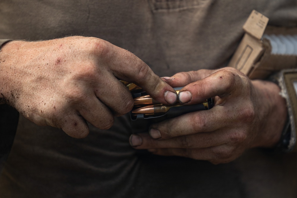 A close-up photograph of a pair of weathered, dirty hands loading ammunition into a black firearm magazine. The hands appear calloused and work-worn, suggesting an experienced individual. Several brass-cased cartridges with copper-jacketed bullets are visible being handled.
