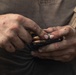 A close-up photograph of a pair of weathered, dirty hands loading ammunition into a black firearm magazine. The hands appear calloused and work-worn, suggesting an experienced individual. Several brass-cased cartridges with copper-jacketed bullets are visible being handled.