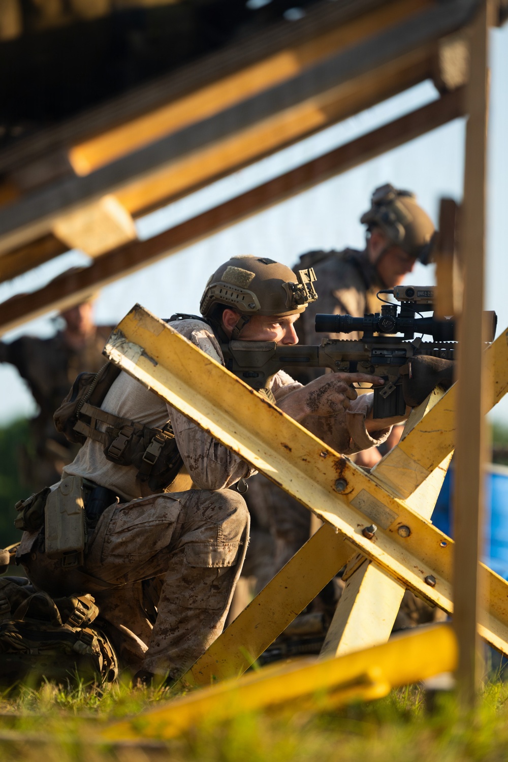 A military service member in desert camouflage (MARPAT pattern) is crouched behind yellow wooden barriers, aiming a rifle equipped with an optical scope. At least one additional service member is visible in the background in similar gear. The setting appears to be a training exercise or tactical drill, likely at a military range or simulated urban environment.