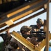 A military service member in desert camouflage (MARPAT pattern) is crouched behind yellow wooden barriers, aiming a rifle equipped with an optical scope. At least one additional service member is visible in the background in similar gear. The setting appears to be a training exercise or tactical drill, likely at a military range or simulated urban environment.